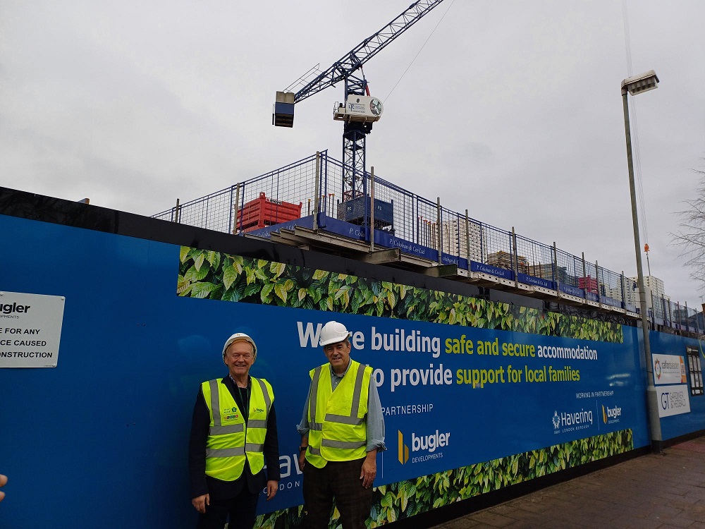 Image shows two men smiling to the camera standing in front of a tall hoarding with a crane in the background